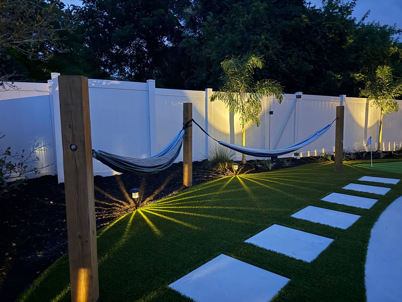 Two hammocks strung between posts along the fence at dusk, path lights fanning across the turf.