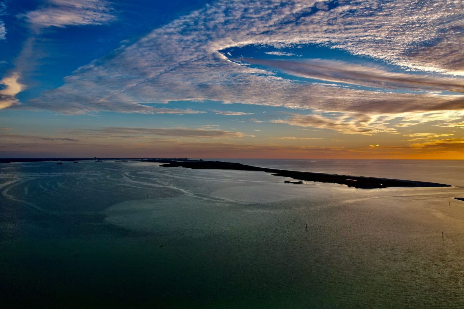 An aerial over the Gulf at sunset: a barrier island lies dark against an orange horizon.